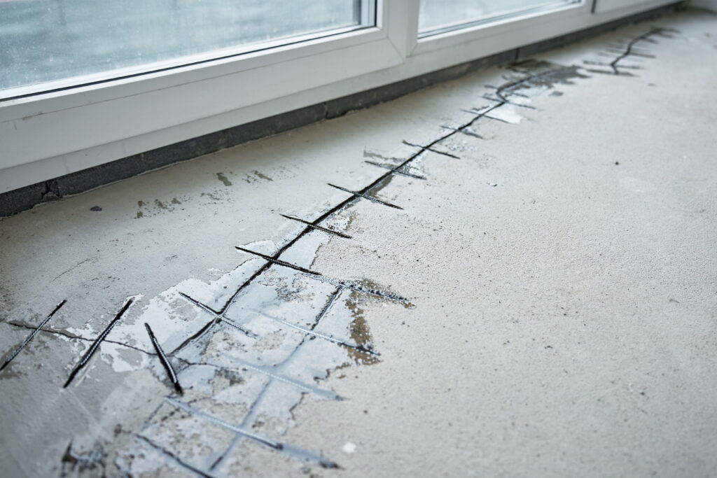 Concrete floor crack reinforced with rebar stitching staples along basement window base showing structural leak repair