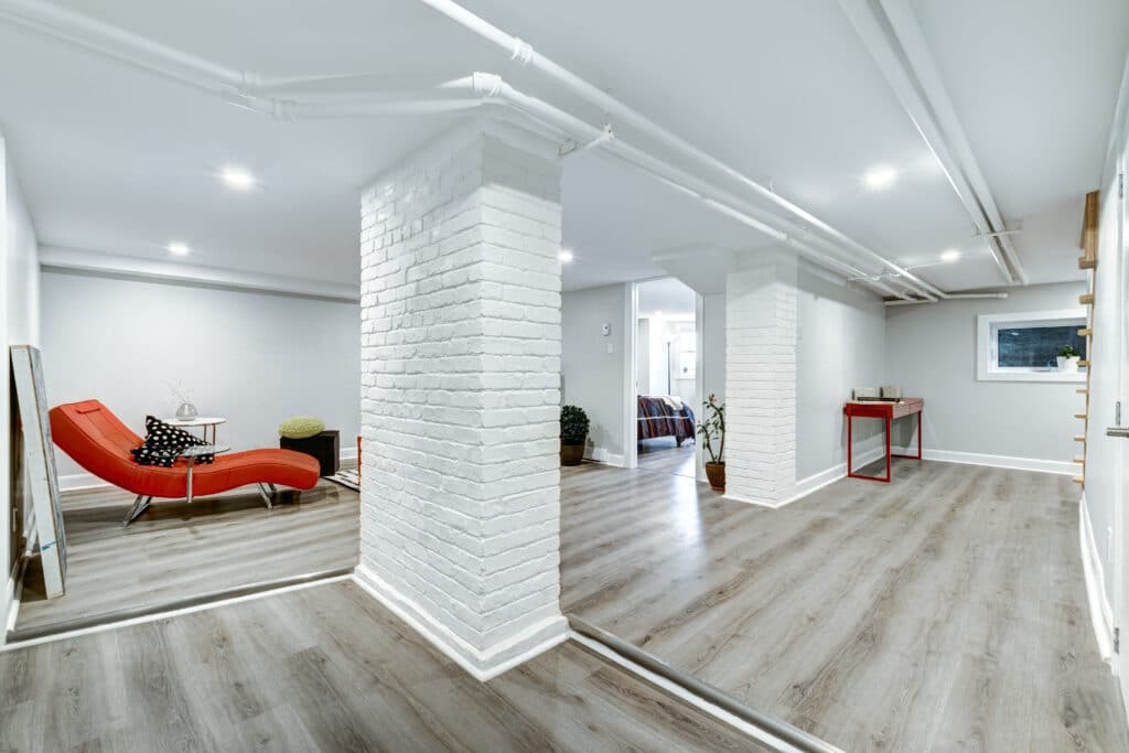 Renovated basement with laminate flooring, painted white brick columns, exposed pipe ceiling and open layout living area