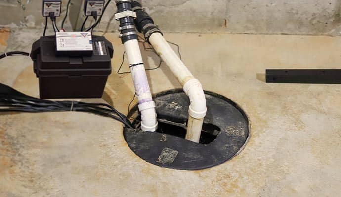Sump pump pit with sealed cover, dual PVC discharge pipes and battery backup unit on basement concrete floor